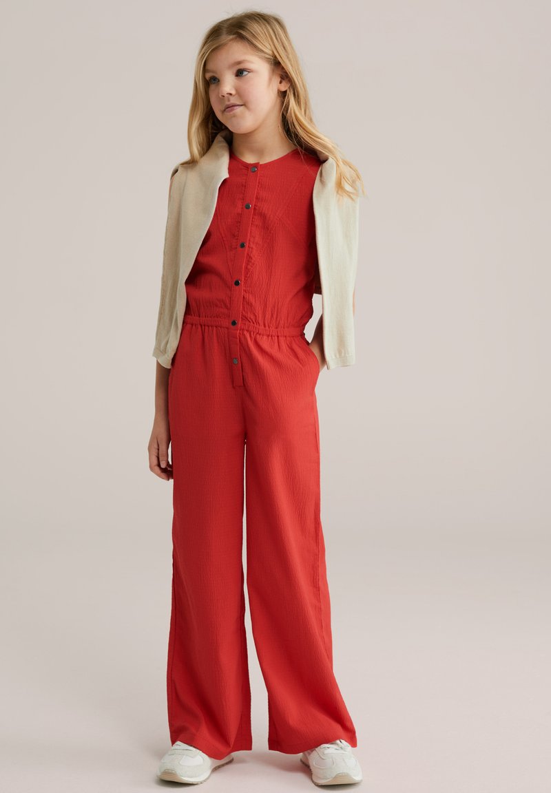 Combinaison rouge à manches courtes, fermeture à boutons et taille élastique. Associée à un cardigan beige clair. Des baskets blanches complètent la tenue.