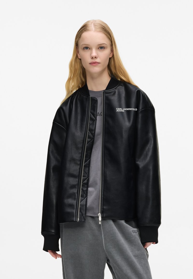 Νεαρή γυναίκα με μακριά ξα�νθά μαλλιά, φορώντας μια μαύρη δερμάτινη τζάκετ με φερμουάρ και γκρίζες casual παντελόνες μπροστά από άσπρο φόντο.