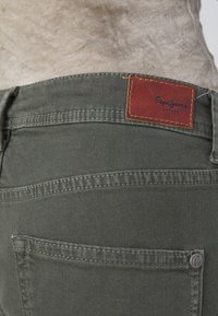 Jeans de mezclilla verde oscuro, con una etiqueta de cuero marrón que lleva el logo de "Pepe Jeans", costuras detalladas y un bolsillo trasero.