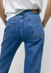 Korkeavyötäröiset siniset farkut, joissa on hieman haalistunut pinta, kahdella takataskulla ja ruskealla nahkakorella vyötäröltä.