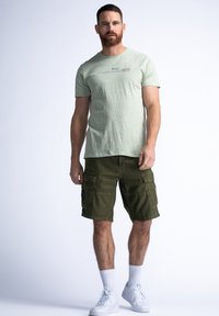 Light green short-sleeve t-shirt with text, paired with olive green cargo shorts featuring multiple pockets and a patch design. White sneakers and socks.