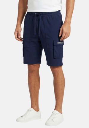 Homme portant un short cargo bleu marine avec taille à cordon de serrage et des baskets blanches, debout devant un fond uni.