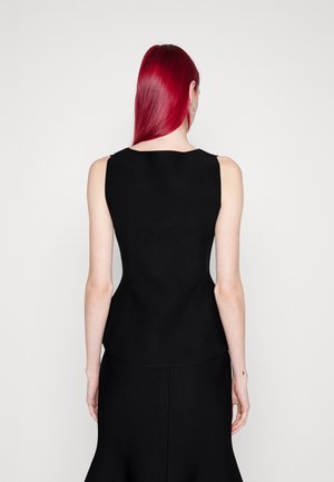 Pitkähiuksinen punatukkainen henkilö pukeutuneena istuvaan mustaan hihattomaan toppiin ja mustaan hameeseen, esitettyinä selästä yksinkertaisen valkoisen taustan edessä.