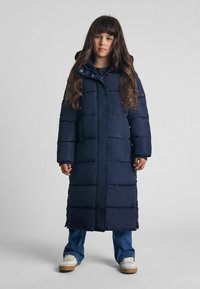 Námornícky puffer kabát s kapucňou, s horizontálnym prešívaním, dlhým strihom a zapínaním na cvoky. V kombinácii s džínsami a teniskami.