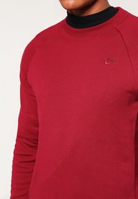 Röd Nike-tröja med rund hals, långa raglanärmar, ribbade muddar och en liten broderad logotyp på bröstet. Slät textur.