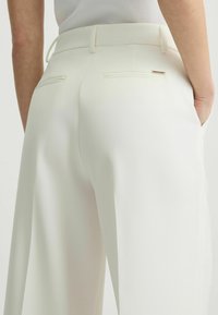 Witte op maat gemaakte broek met een gladde stof, slim fit, dubbele achterzakken en een subtiele gouden accent nabij één zak.