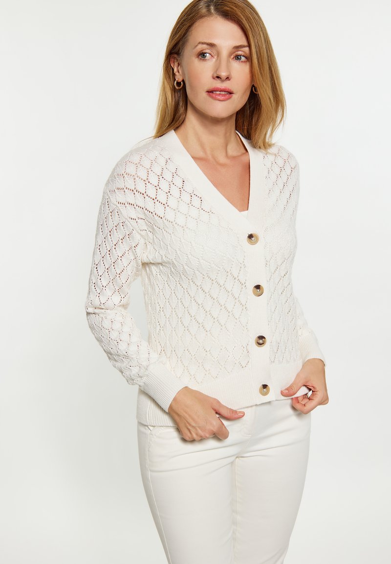 Cardigan alb tricotat cu model în diamant, decolteu în V, mâneci lungi și nasturi mari bej. Materialul pare moale și texturat.