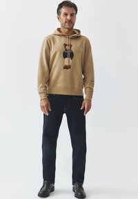 Sudadera beige con un gráfico de oso en la parte delantera, combinada con jeans rectos de color azul oscuro y zapatos negros. Diseño simple y casual.