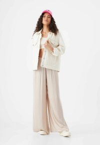 Weiße Denimjacke über einem hellbeigen Crop-Top, kombiniert mit weit geschnittenen beigen Hosen und cremefarbenen Slippern. Helles pinkes Cap auf dem Kopf.