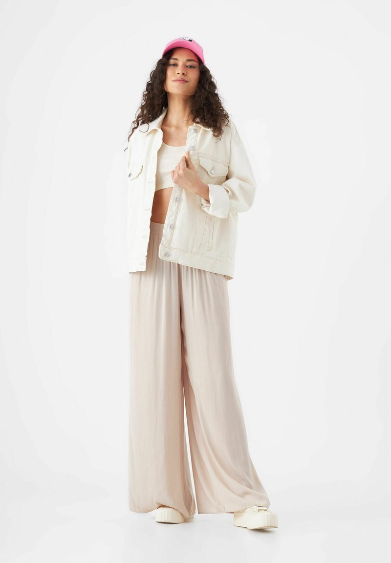 Weiße Denimjacke über einem hellbeigen Crop-Top, kombiniert mit weit geschnittenen beigen Hosen und cremefarbenen Slippern. Helles pinkes Cap auf dem Kopf.