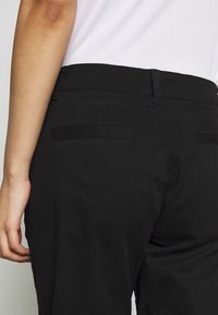 Côté gauche d'une personne portant un pantalon noir et une chemise blanche, main détendue sur le côté, poches arrière visibles sur le pantalon.