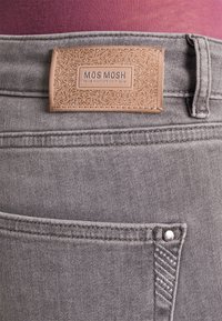 Grijze denim jeans achterzak met decoratieve stiksels en een tan leren merklabel met de tekst "MOS MOSH, WE ARE PASSIONATE ABOUT JEANS".