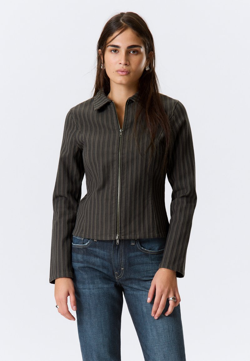 Donkergroene gestreepte jas van lichte stof, met een ritssluiting en puntige kraag, gecombineerd met blauwe jeans.