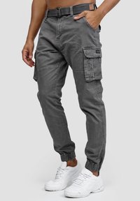 Pantalon cargo gris avec poignets élastiques, multiples poches et coupe décontractée. Porté avec des baskets blanches. Confectionné dans un tissu doux et durable.
