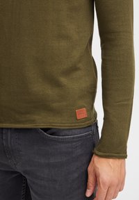 Olijfgroene shirt met lange mouwen en een gestructureerd breipatroon. Heeft een bruine leren logo patch nabij de zoom, gecombineerd met donkergrijze broek.