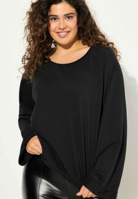 Top nero a maniche lunghe con scollo tondo e vestibilità comoda, realizzato in tessuto morbido con dettagli di cucitura visibili, abbinato a leggings neri lucidi.