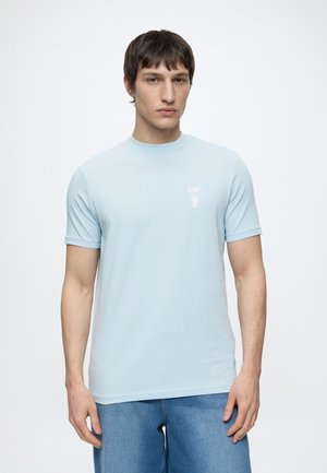 Jeune homme aux cheveux courts et foncés, portant un t-shirt bleu clair à manches courtes et un jean bleu sur un fond blanc uni.