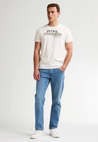 Męski kremowy t-shirt z napisem "PETROL INDUSTRIES & CO" w zielonym i czarnym kolorze; zestawiony z jasnoniebieskimi dżinsami i białymi tenisówkami.