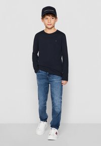Poiss kannab tumesinist pika varrukaga särki, siniseid teksaseid ja valgeid tossusid. Must müts, millel on valge tekst "TOMMY HILFIGER".