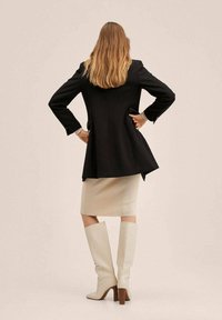 Femme aux longs cheveux blonds portant un blazer noir, une jupe crayon beige et des bottes à talons beiges montantes, se tenant les mains sur les hanches, de dos.