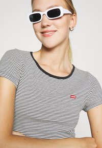 Tricou cu dungi alb-negru, cu mânecă scurtă, guler rotund și un mic logo Levi's roșu. Accompanied by ochelari de soare pătrați albi.
