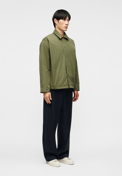 Νεαρός άνδρας στέκεται φορώντας ένα χακί πράσινο σακάκι με κουμπιά, μαύρο παντελόνι φαρδύ με κάθετες ρίγες και λευκά αθλητικά παπούτσια, σε απλό φόντο.