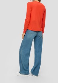 Röd långärmad topp i kombination med ljusblå vida jeans. Toppen har en rund halsringning; jeansen har detaljerade bakfickor. Vita sneakers.