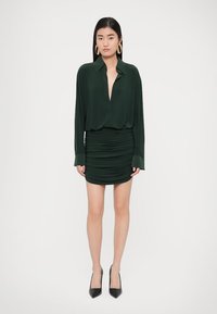 Naine, kes kannab tumerohelist pika varrukaga pluusi ja kangutatud miniseelikut, mustad teravnurksed kontsakingad ning suured rõngaskõrvarõngad, seisab valge tausta ees.