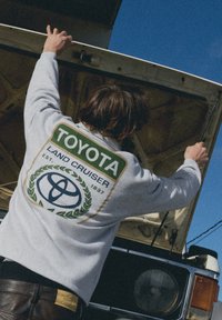 Felpa grigia con una grande grafica verde e oro con la scritta "TOYOTA LAND CRUISER" e un logo circolare sul retro.