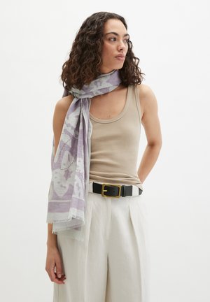 Femme aux cheveux bouclés portant un débardeur beige, un pantalon blanc avec une ceinture noire, et une écharpe à motifs lilas drapée sur une épaule.