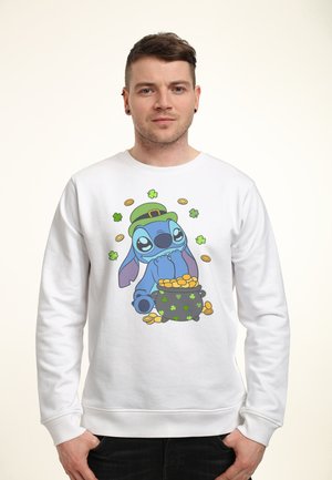 Man met een witte trui met een cartoon van een blauwe alien die een groene leprechaun-hoed draagt, naast een pot met gouden munten en vliegende klavertjes vier.