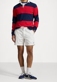 Muž oblečený v námořnicky modrém a červeném pruhovaném ragbyovém tričku, bílých šortkách na šňůrku, bílých ponožkách a námořnicky modrých teniskách, stojící s jednou rukou v kapse.