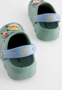 Ljusblå crocs med en mintgrön kropp, prydda med dekorationer av tecknade figurer och en rem som är märkt "PAW PATROL." Texturerad yta och ventilerande hål finns.