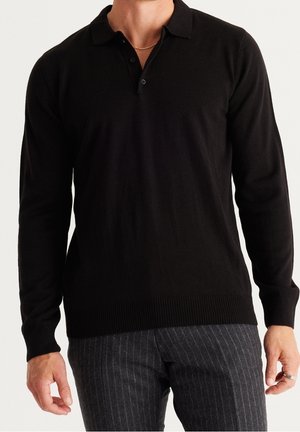 Homme portant un polo noir à manches longues et un pantalon gris à fines rayures, avec une chaîne en argent et une bague à la main droite.