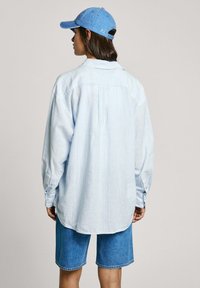 Hellblau gestreifte, knöpfbare Bluse mit langen Ärmeln, kombiniert mit Jeansshorts. Die Bluse hat eine lässige Passform und einen abgerundeten Saum.