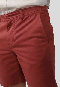 Shorts rouges en coton avec une texture lisse, dotés de poches latérales et de détails cousus. La taille comporte un cordon de serrage pour un ajustement.