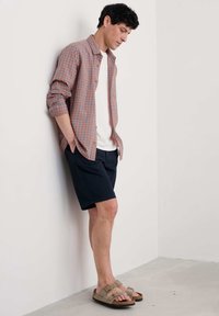 Tjekket skjorte i orange og blå, hvid T-shirt under, marineblå shorts og brune slide-sandaler med to stropper, stående op ad en væg.