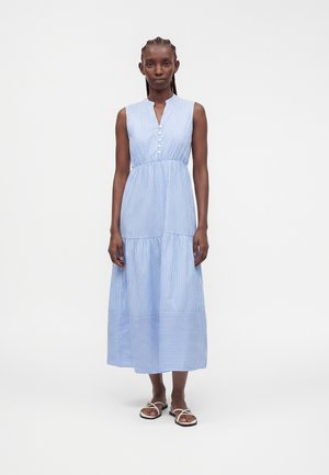 Femme debout portant une robe midi sans manches à rayures verticales bleues et blanches avec des boutons et des sandales plates blanches sur fond blanc.