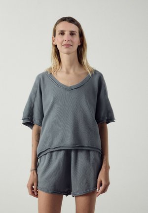 Femme aux cheveux mi-longs, portant un t-shirt ample gris à col en V et un short assorti, debout devant un fond clair uni.