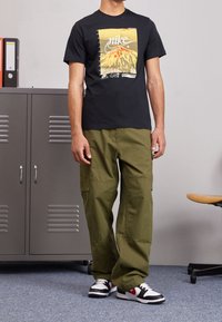 Svart t-shirt med grafiskt tryck, olivgröna cargobyxor och svarta sneakers med röda och vita detaljer, mot en neutral bakgrund.