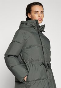 Giacca anorak imbottita di colore verde scuro con chiusura a zip, cappuccio regolabile e vita elastica per un look aderente. Presenta tasche laterali.