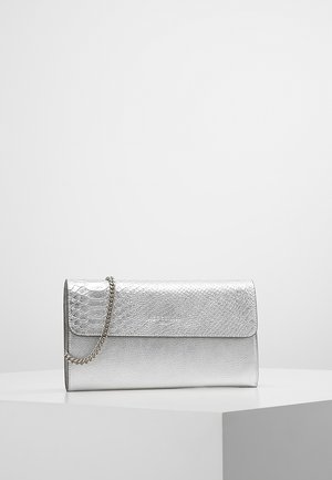 Pochette argentée texturée avec une bandoulière chaîne, placée sur une surface géométrique blanche sur un fond gris uni.