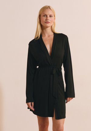 Blonde Frau, die ein schwarzes, langärmeliges Wickelkleid trägt, das an der Taille gebunden ist, stehend vor einem schlichten beigen Hintergrund.