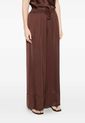 Donna con pantaloni palazzo larghi in raso marrone con vita elasticizzata e coulisse, abbinati a sandali infradito neri.