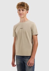 Tienerjongen met lichtbruin haar, gekleed in een beige T-shirt met de tekst "BALLIN'" en blauwe jeans, die staat tegen een effen lichte achtergrond.