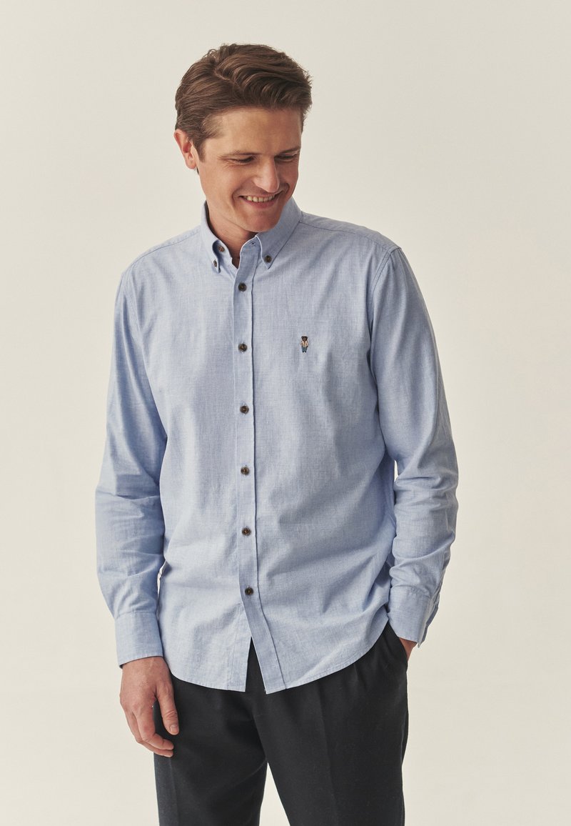 Cămașă bleu deschis, cu butoni maroni și manșete lungi, confecționată dintr-un material texturat, având un mic logo brodat.