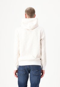 Light beige hoodie with patterned texture, front pockets, and a drawstring hood, worn with dark blue jeans, viewed from the back.