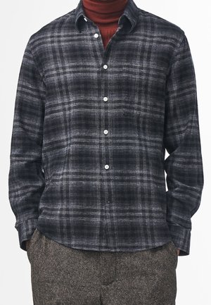 Mann trägt ein schwarz-grau kariertes Hemd über einem rostfarbenen Rollkragenpullover, kombiniert mit braunen, strukturierten Hosen, Hände in den Taschen.