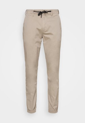 Beige verjüngte Baumwollhose mit elastischem Bund und schwarzem Kordelzug. Verfügt über Seitentaschen und elastische Bündchen an den Knöcheln.