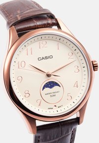 Pyöreä Casio-kello, jossa on kerma-nuotti, ruusukultaiset viisarit ja numerot sekä ruskea nahkahihna. Ominaisuuksiin kuuluu 50 metrin vesitiiviys.